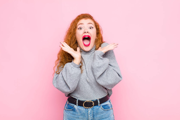 young red head woman feeling shocked and excited, laughing, amazed and happy because of an unexpected surprise against pink wall - Photo, image