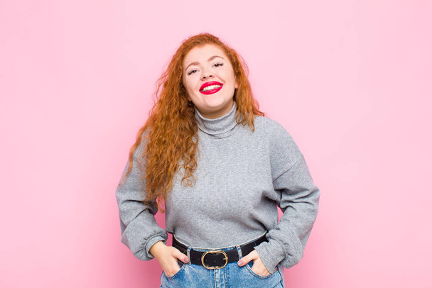 young red head woman smiling cheerfully and casually with a positive, happy, confident and relaxed expression against pink wall - Foto, afbeelding
