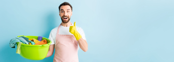 Panoramaaufnahme eines glücklichen Mannes, der eine Plastikwaschschschüssel mit schmutziger Wäsche in der Hand hält und den Daumen auf blau zeigt  - Foto, Bild