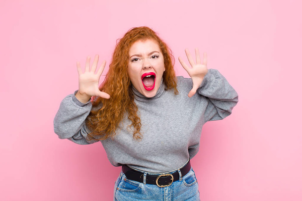 young red head woman screaming in panic or anger, shocked, terrified or furious, with hands next to head against pink wall - Photo, image