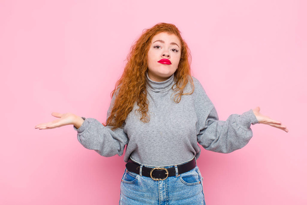 young red head woman feeling puzzled and confused, unsure about the correct answer or decision, trying to make a choice against pink wall - Fotoğraf, Görsel