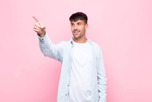 young handsome man feeling shocked and surprised, pointing and looking upwards in awe with amazed, open-mouthed look against pink wall - Foto, imagen