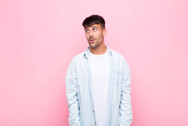 young handsome man feeling shocked, happy, amazed and surprised, looking to the side with open mouth against pink wall - 写真・画像