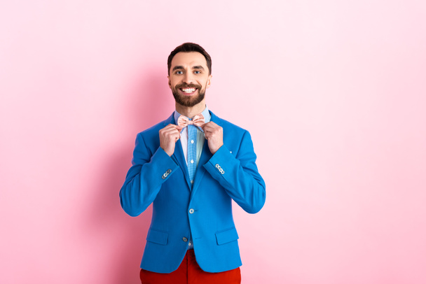 happy bearded man in suit touching bow tie on pink  - Фото, изображение