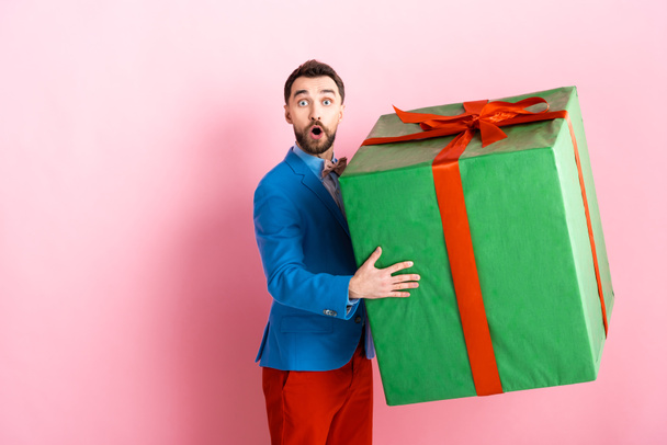 удивленный бородатый мужчина держит огромную подарочную коробку на розовом фоне
  - Фото, изображение