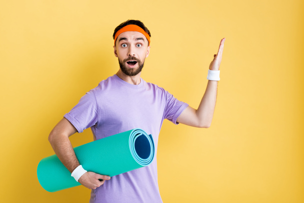 surprised sportsman holding fitness mat and pointing with hand isolated on yellow - Photo, Image