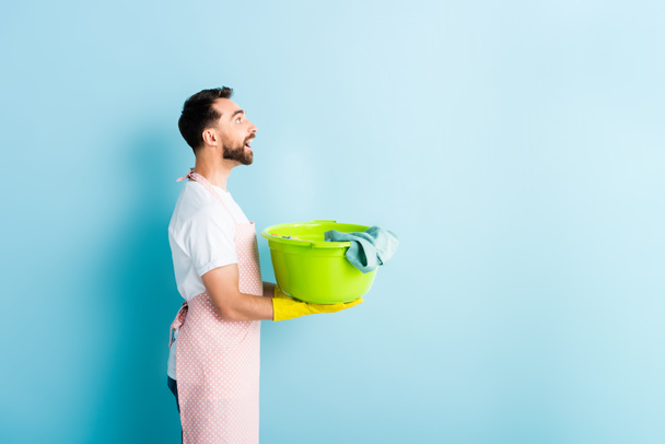 Seitenansicht eines glücklichen bärtigen Mannes, der schmutzige Wäsche auf blau hält  - Foto, Bild