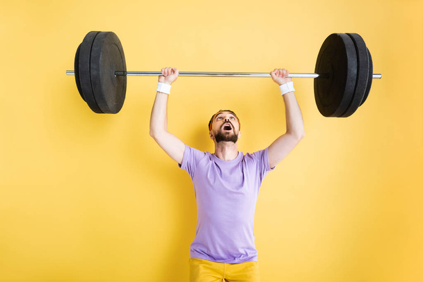 surprised bearded sportsman looking at barbell while working out on yellow - Foto, Bild