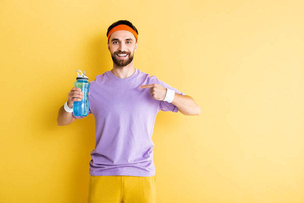 bearded sportsman pointing with finger at sports bottle on yellow - Фото, изображение