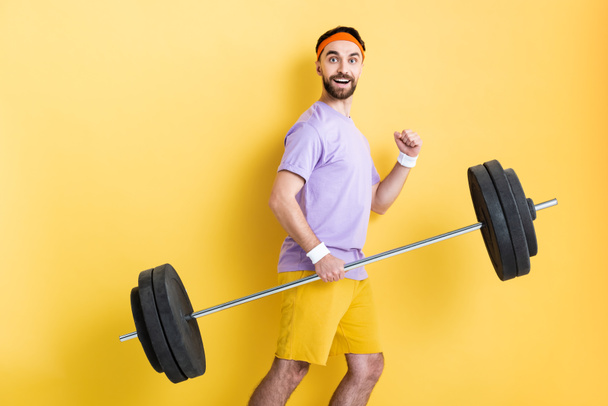 щасливий спортсмен тримає важкий штангу під час ходьби на жовтому
 - Фото, зображення