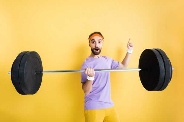 surprised sportsman pointing with finger and holding barbell on yellow - Photo, Image