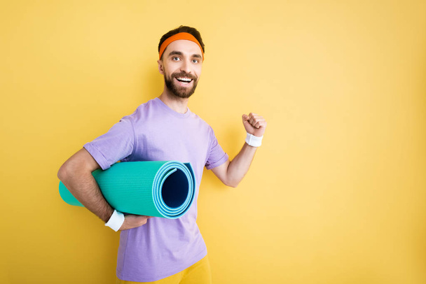 cheerful bearded sportsman holding fitness mat on yellow - Photo, Image