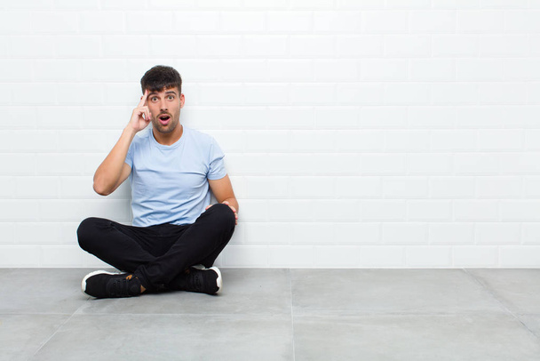 young handsome man looking surprised, open-mouthed, shocked, realizing a new thought, idea or concept sitting on cement floor - Фото, изображение