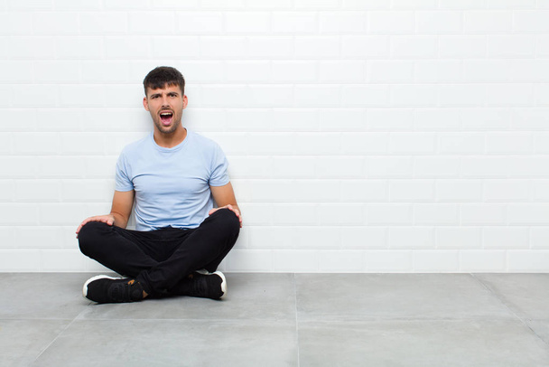 young handsome man feeling terrified and shocked, with mouth wide open in surprise sitting on cement floor - Photo, image