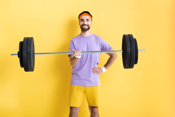 cheerful sportsman standing with hand on hip and holding barbell on yellow - Фото, зображення