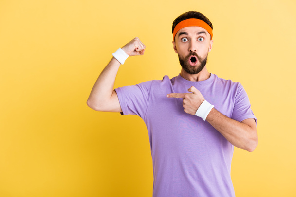 shocked man in headband pointing with finger and muscle isolated on yellow - Фото, изображение