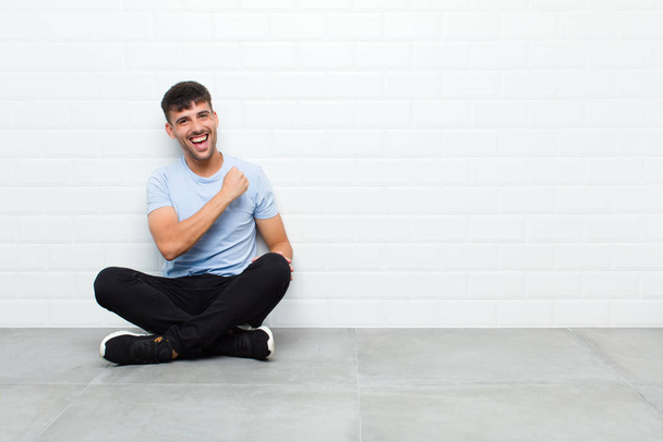 young handsome man feeling happy, positive and successful, motivated when facing a challenge or celebrating good results sitting on cement floor - Фото, зображення