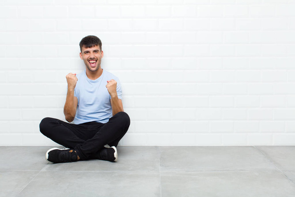 young handsome man shouting triumphantly, laughing and feeling happy and excited while celebrating success sitting on cement floor - Foto, Imagen