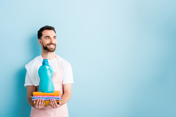 homme barbu heureux tenant des vêtements propres et détergent à lessive sur bleu
  - Photo, image