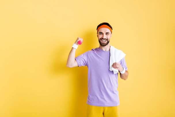cheerful sportsman with dumbbell holding towel on yellow - Photo, Image