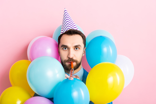 hombre barbudo en gorra de fiesta con soplador de fiesta en la boca cerca de globos en rosa
  - Foto, Imagen