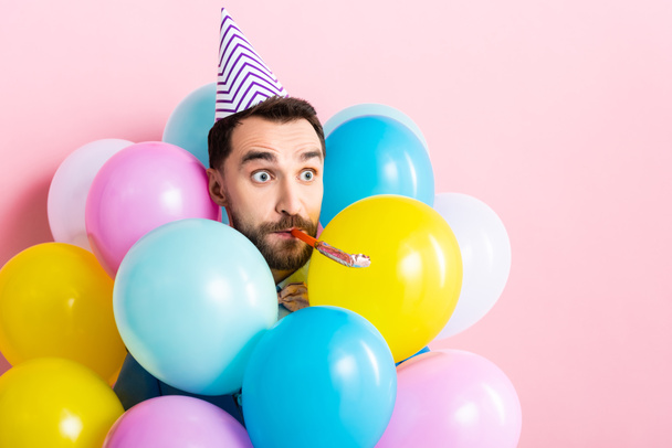handsome man in party cap with party blower in mouth near balloons on pink  - Foto, afbeelding