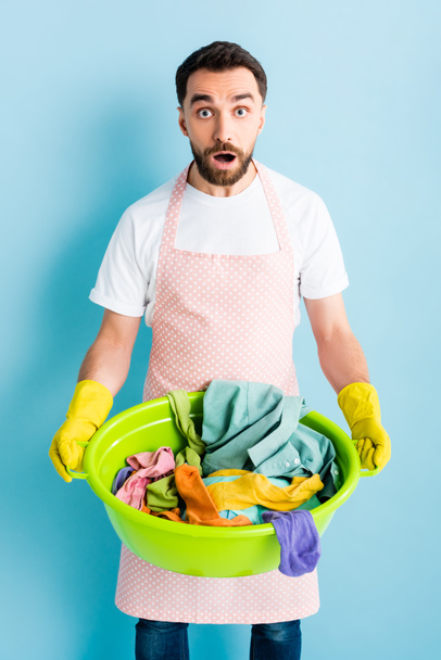 homme choqué dans des gants en caoutchouc tenant bol de lavage avec buanderie sale sur bleu
  - Photo, image
