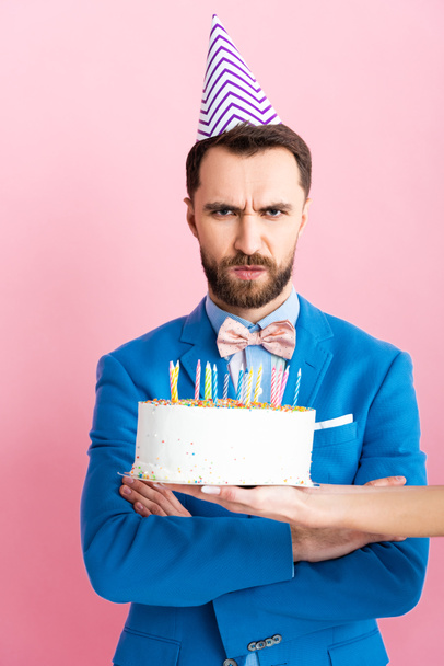 vista ritagliata di donna che tiene torta di compleanno vicino uomo offeso con braccia incrociate isolato su rosa
  - Foto, immagini
