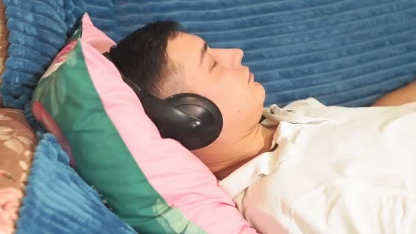 Genç adam evdeki kulaklıklarla kanepede dinleniyor. Rahatlatıcı müzik dinliyor, sakin müzik dinliyor. Rahatlama, gönül rahatlığı. Kulaklıklar ve müzik dinlemek. - Video, Çekim