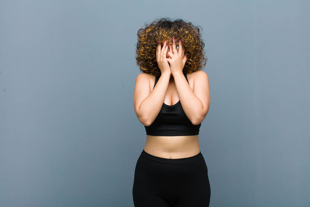 young sports woman covering face with hands, peeking between fingers with surprised expression and looking to the side against gray wall - Foto, Imagem
