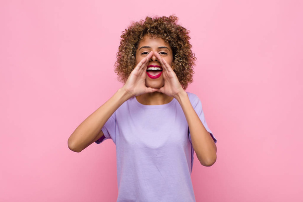 young african american woman feeling happy, excited and positive, giving a big shout out with hands next to mouth, calling out against pink wall - Φωτογραφία, εικόνα