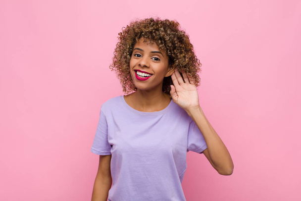 young african american woman smiling, looking curiously to the side, trying to listen to gossip or overhearing a secret against pink wall - Photo, Image
