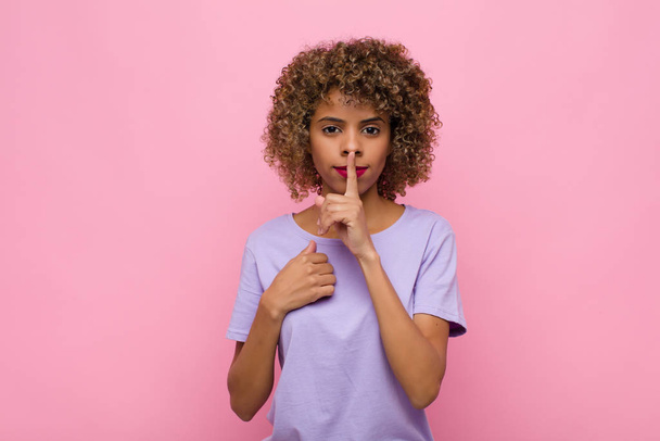 young african american woman looking serious and cross with finger pressed to lips demanding silence or quiet, keeping a secret against pink wall - Photo, Image