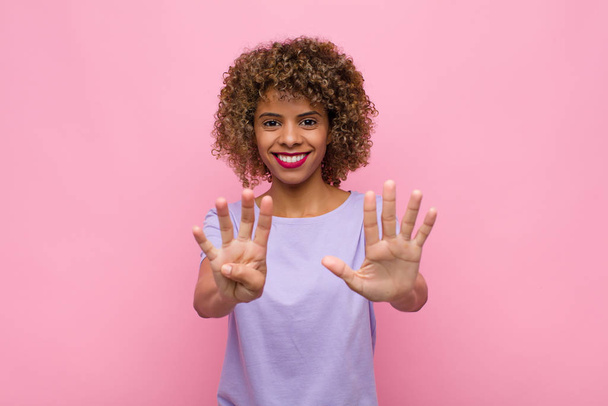 młoda Afroamerykanka uśmiecha się i wygląda przyjaźnie, pokazuje numer dziewięć lub dziewiąty z ręką do przodu, odliczając w dół od różowej ściany - Zdjęcie, obraz