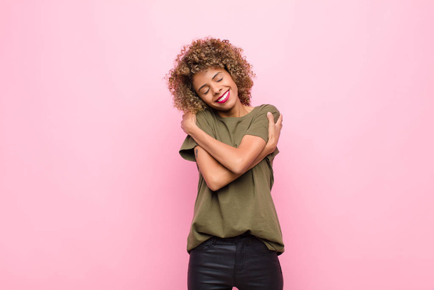 young african american woman feeling in love, smiling, cuddling and hugging self, staying single, being selfish and egocentric against pink wall - Фото, изображение
