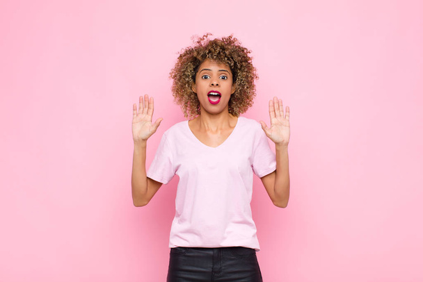 young african american woman feeling stupefied and scared, fearing something frightening, with hands open up front saying stay away against pink wall - 写真・画像