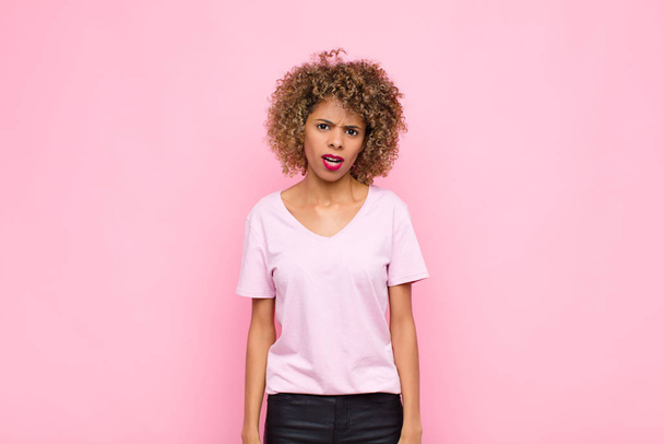 young african american woman feeling puzzled and confused, with a dumb, stunned expression looking at something unexpected against pink wall - 写真・画像
