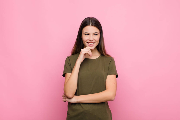 young pretty woman smiling with a happy, confident expression with hand on chin, wondering and looking to the side against pink wall - Photo, image