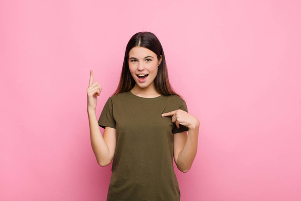 young pretty woman feeling proud and surprised, pointing to self confidently, feeling like successful number one against pink wall - Fotoğraf, Görsel