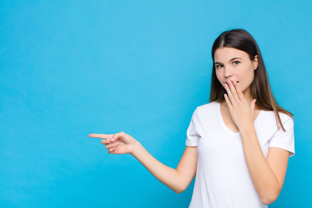 young pretty woman feeling happy, shocked and surprised, covering mouth with hand and pointing to lateral copy space against blue wall - Foto, Imagen