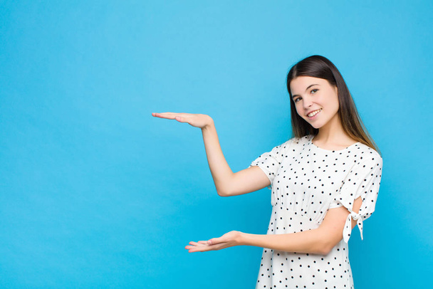 young pretty woman smiling, feeling happy, positive and satisfied, holding or showing object or concept on copy space against blue wall - Fotoğraf, Görsel