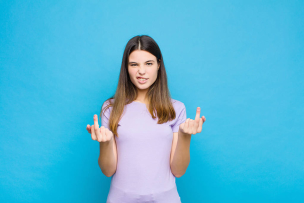young pretty woman feeling provocative, aggressive and obscene, flipping the middle finger, with a rebellious attitude against blue wall - 写真・画像