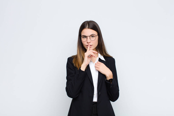 young businesswoman looking serious and cross with finger pressed to lips demanding silence or quiet, keeping a secret against white wall - Photo, image