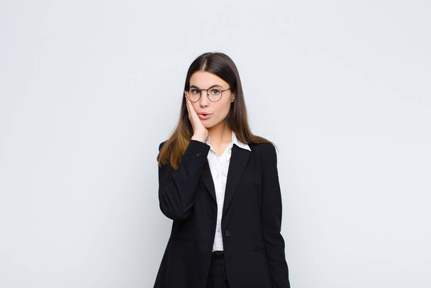 young businesswoman feeling shocked and astonished holding face to hand in disbelief with mouth wide open against white wall - Fotografie, Obrázek