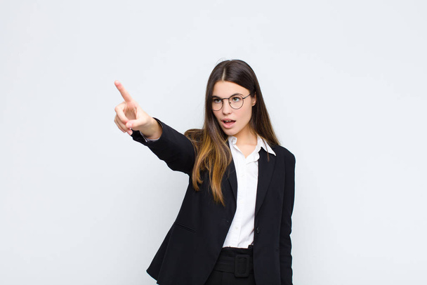 young businesswoman feeling shocked and surprised, pointing and looking upwards in awe with amazed, open-mouthed look against white wall - Foto, afbeelding
