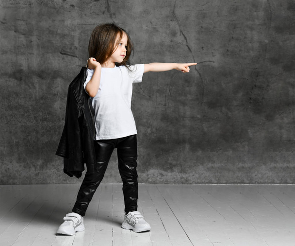 Menina pequena em preto e branco rock star estilo casual roupas e tênis brancos de pé e apontando para o lado com o dedo
 - Foto, Imagem