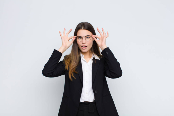 young businesswoman feeling shocked, amazed and surprised, holding glasses with astonished, disbelieving look against white wall - Photo, image