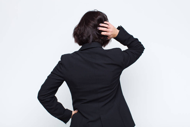 young businesswoman thinking or doubting, scratching head, feeling puzzled and confused, back or rear view against white wall - Foto, afbeelding
