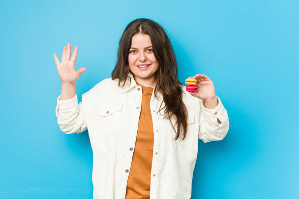 Jovem mulher curvilínea segurando macaroons sorrindo alegre mostrando número cinco com os dedos
. - Foto, Imagem