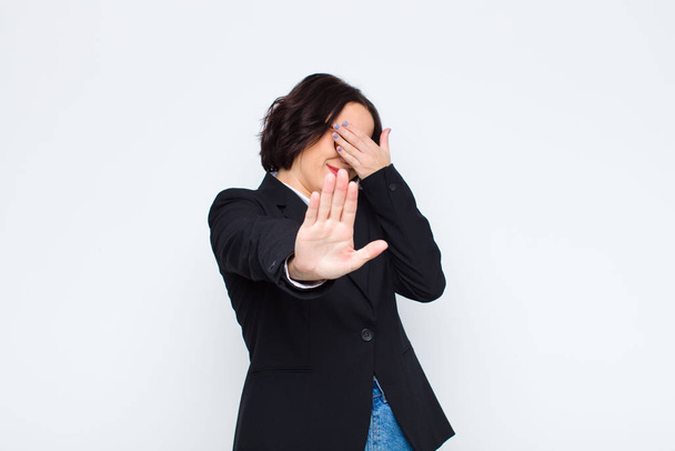 young businesswoman covering face with hand and putting other hand up front to stop camera, refusing photos or pictures against white wall - Foto, afbeelding